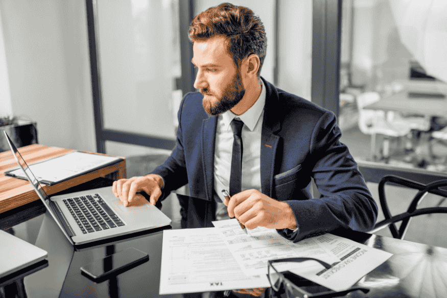 Homme en costume analysant des documents financiers à son bureau avec un ordinateur portable.